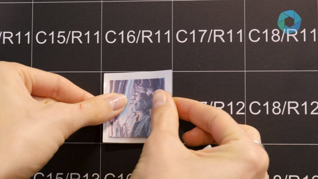 A person's hands place a small, partially peeled photo onto a black grid with white alphanumeric labels like "C15/R11" and "C16/R11." A blue geometric logo appears in the top right, perfect for a corporate event experiential activation.