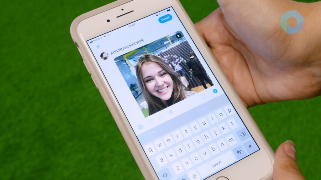 A hand holds a mobile displaying a Twitter post draft with a photo of a smiling woman and the hashtag "#photomosaicw." The bright green background suggests a creative setting, perfect for showcasing sketch bot hire or draw me bot hire at corporate event experiential activities.