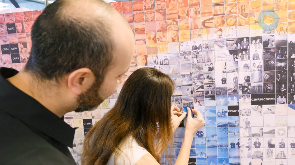 A woman pins a photo onto a colourful wall collage while a man watches her. The collage, created with premium photo booth hire, is made up of many small, square photos arranged in a grid pattern.