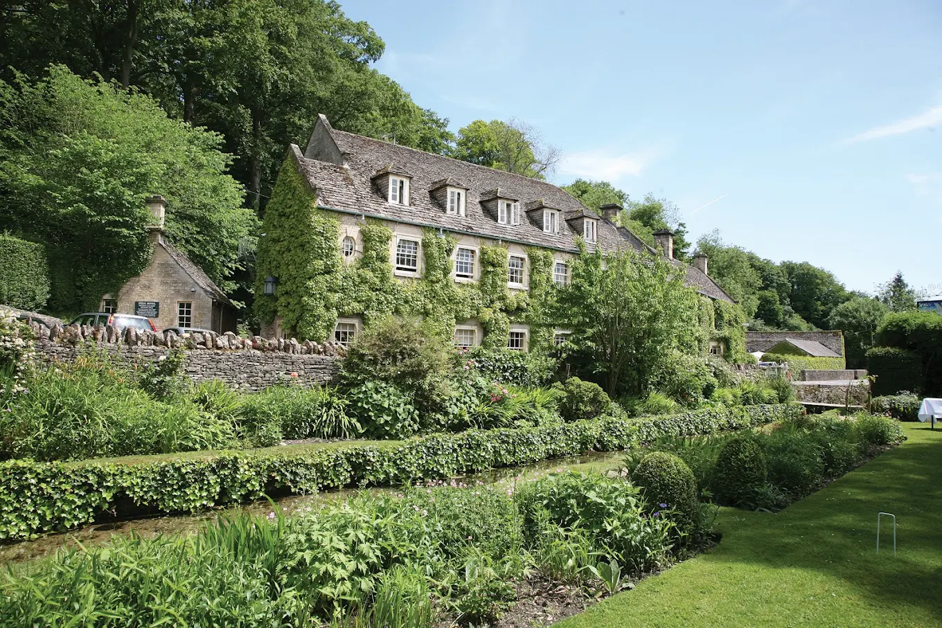 A large stone house covered in ivy sits among lush green gardens, hedges, and neatly trimmed lawns, with trees and blue sky in the background—perfectly capturing the charm of the Swan Hotel in Bibury.
