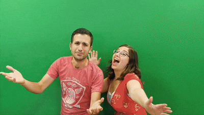 A man and a woman stand in front of a green screen. The man looks confused with his arms out, while the woman enthusiastically gestures and smiles, both wearing red shirts—perfect for showcasing premium photo booth hire.