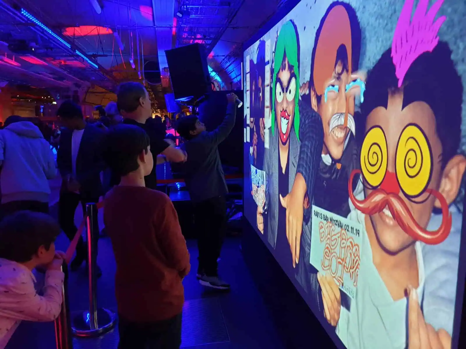 Children watch a large screen displaying their faces with colourful, silly cartoon filters—including exaggerated glasses, moustaches, and hair—in a vibrant, neon-lit indoor space featuring premium photo booth hire.