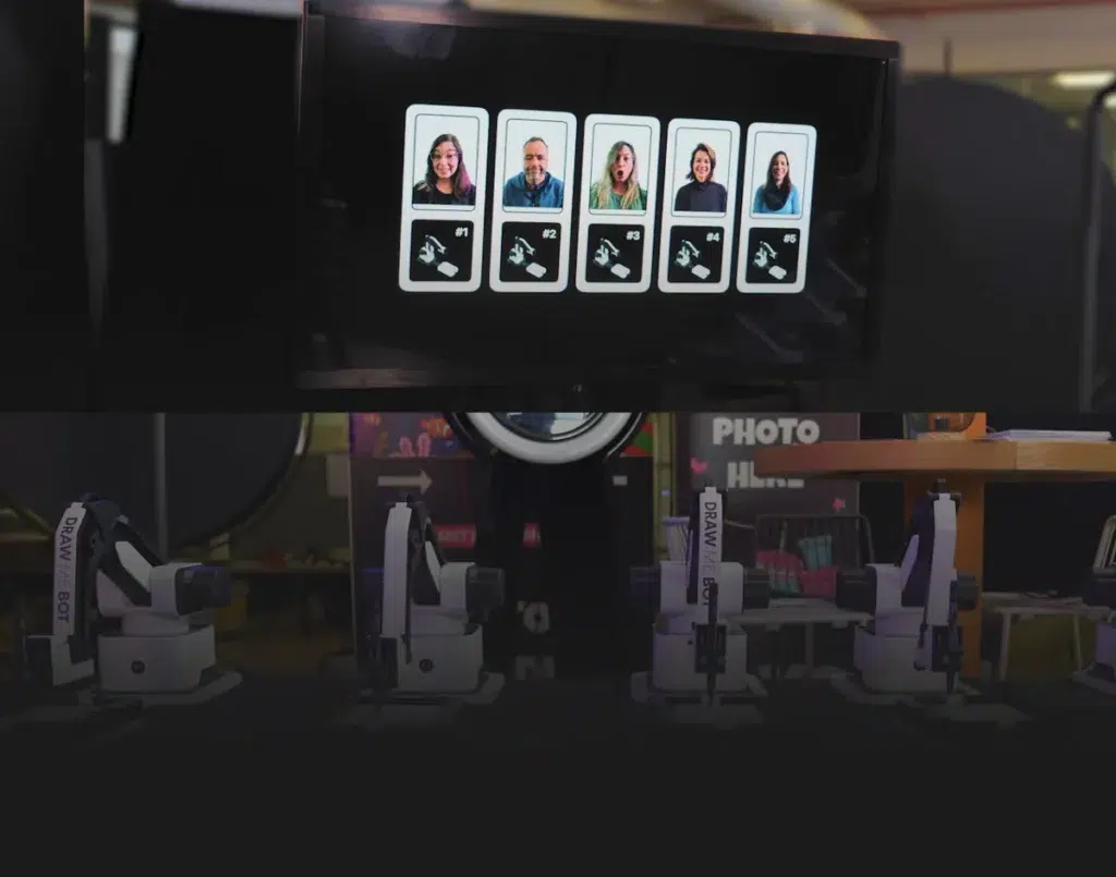 A screen displays five headshots of people, each with information below the photos. Below the screen, several robotic arms labelled "DRAWBOT" are positioned on tables, perfect for corporate event experiential or sketch bot hire experiences.