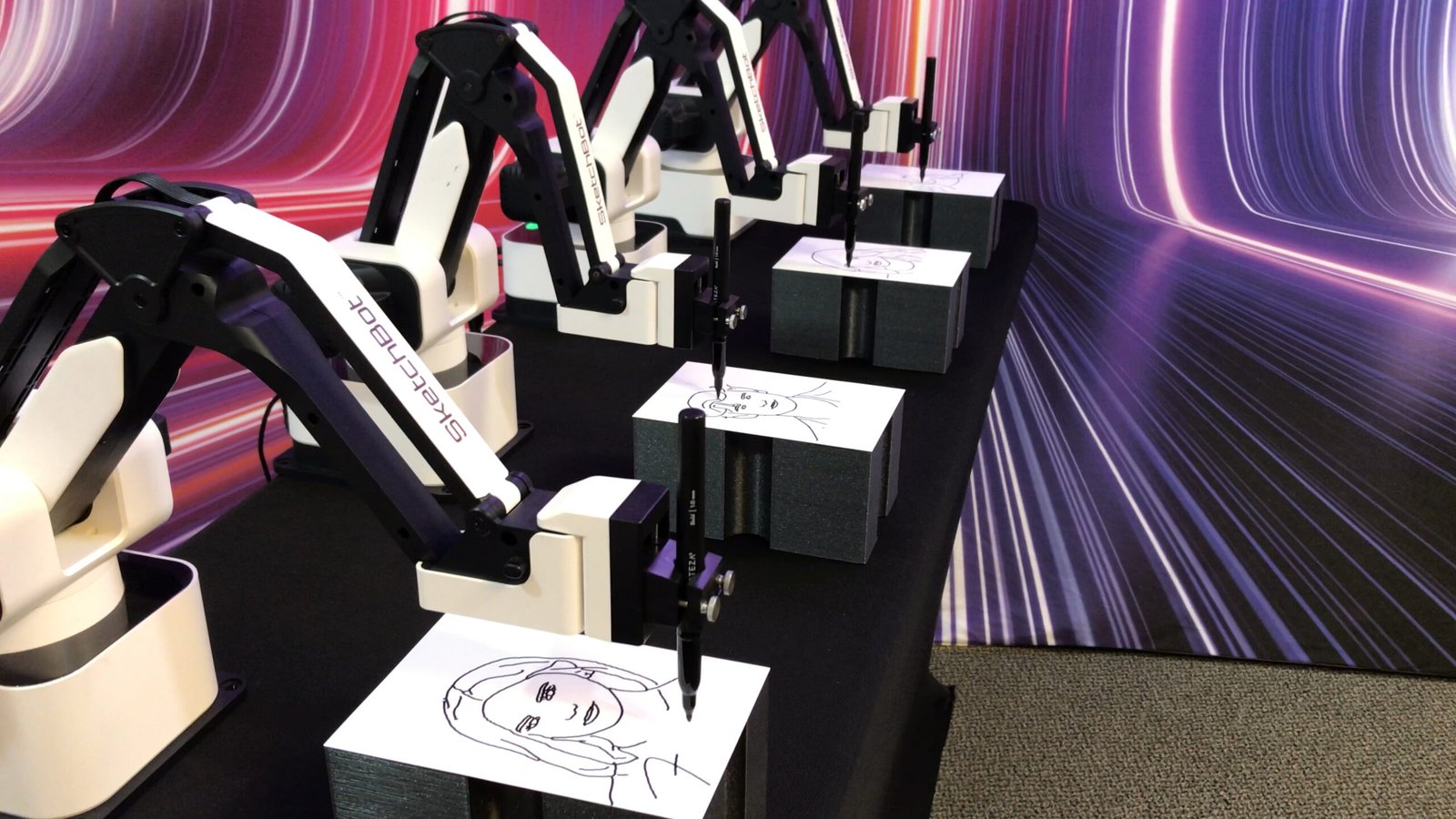 Several robotic arms are drawing portraits on paper, each creating a line art face. The robots are arranged in a row on a black table, with a colourful, futuristic light background—perfect for corporate event experiential or draw me bot hire.