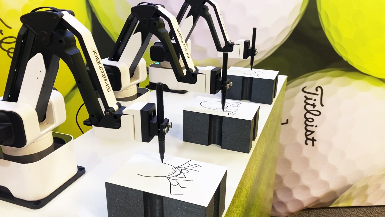 Three robotic arms equipped with pens draw on sheets of paper, each mounted on a foam block. Perfect for corporate event experiential activations, this sketch bot hire set-up is set against large golf balls and a Titleist logo.