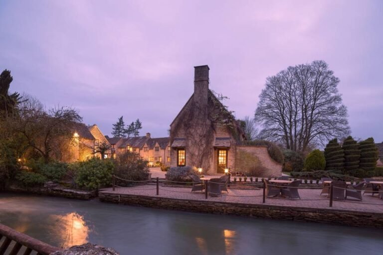 A charming stone cottage with warm lights glowing inside sits beside a calm river at dusk, near the Old Swan and Minster Mill, surrounded by trees and neatly trimmed bushes. Outdoor tables and chairs are arranged by the water.