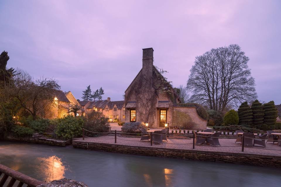 A charming stone cottage with warm lights glowing inside sits beside a calm river at dusk, near the Old Swan and Minster Mill, surrounded by trees and neatly trimmed bushes. Outdoor tables and chairs are arranged by the water.