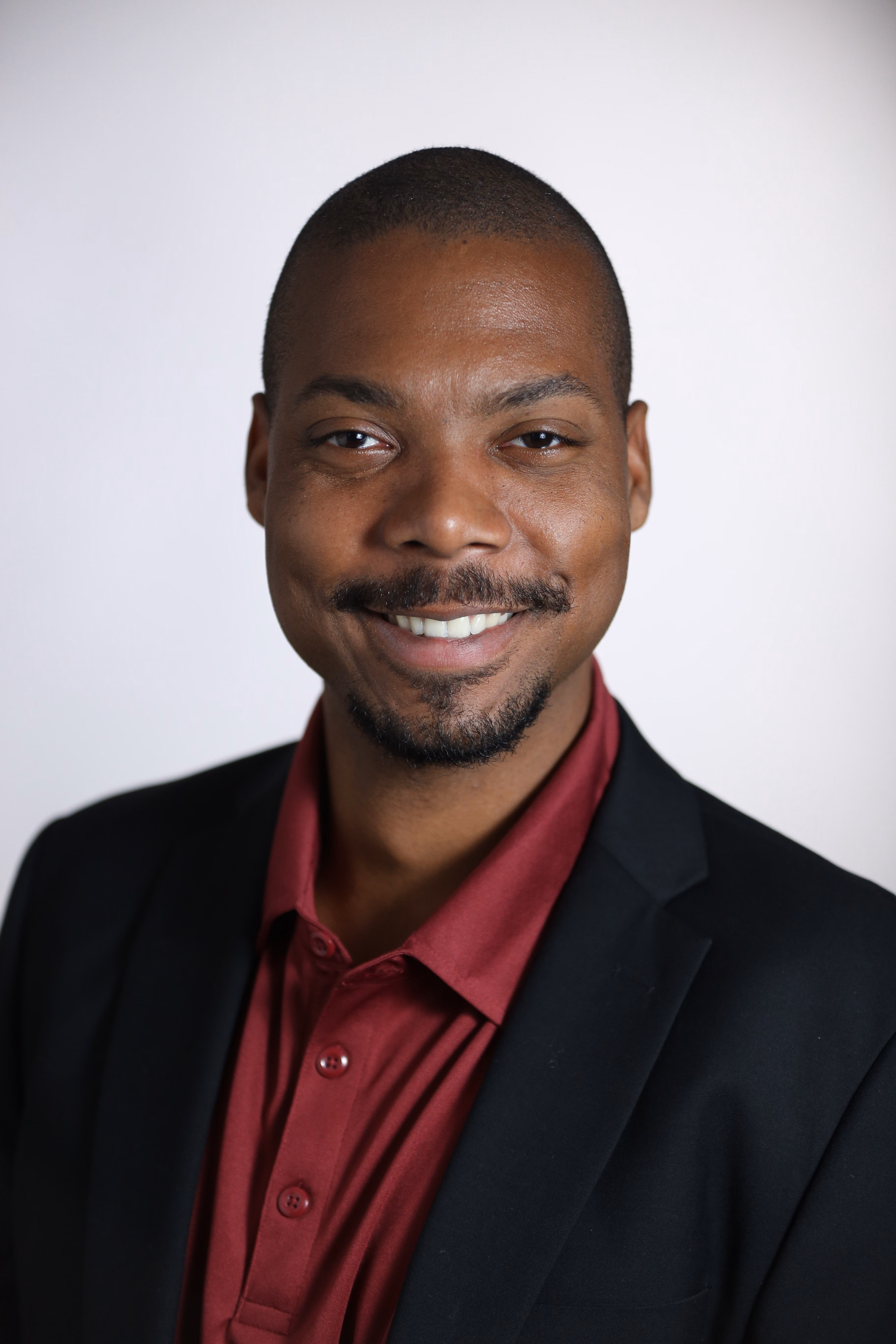 A man in a dark blazer and maroon shirt smiles at the camera against a plain, light background, capturing the engaging spirit of experiential photo marketing.