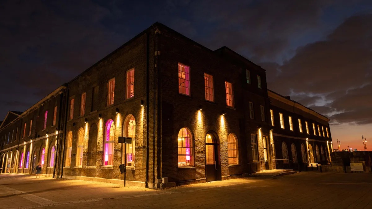A large brick building with arched windows glows with warm yellow and purple lights at dusk, set against a dramatic evening sky—Woolwich Works prepares for the Intuit QuickBooks Event, where Sketchbots bring interactive fun to life.